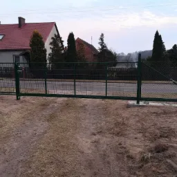 Nowo zamontowana, przesuwna brama wjazdowa w kolorze ciemnej zieleni, widoczna z perspektywy ziemi, z domami w tle i śladami opon na piasku.