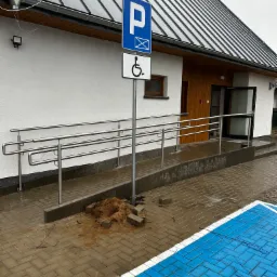 Zewnętrzna rampa dla niepełnosprawnych z nierdzewnymi balustradami prowadząca do budynku z drewnianymi elementami, obok znak parkingu dla niepełnosprawnych, mokra kostka brukowa.