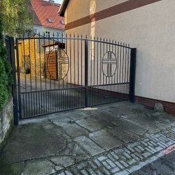 Battig konstrukcje stalowe - Czarna, metalowa brama wjazdowa z symbolem krzyża w okręgu, prowadząca na posesję z budynkiem o beżowej elewacji i czerwonym dachu, widoczna kostka brukowa i fragment zieleni.
