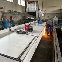 Battig konstrukcje stalowe - Pracownik w warsztacie szlifuje spaw na dużej, metalowej konstrukcji, iskry lecą, widoczny sprzęt spawalniczy i suwnica pod sufitem.