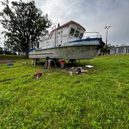 Battig konstrukcje stalowe - Łódź motorowa na metalowych podporach, prace konserwacyjne na trawiastym terenie, widoczne narzędzia i spawarka, pochmurne niebo w tle.