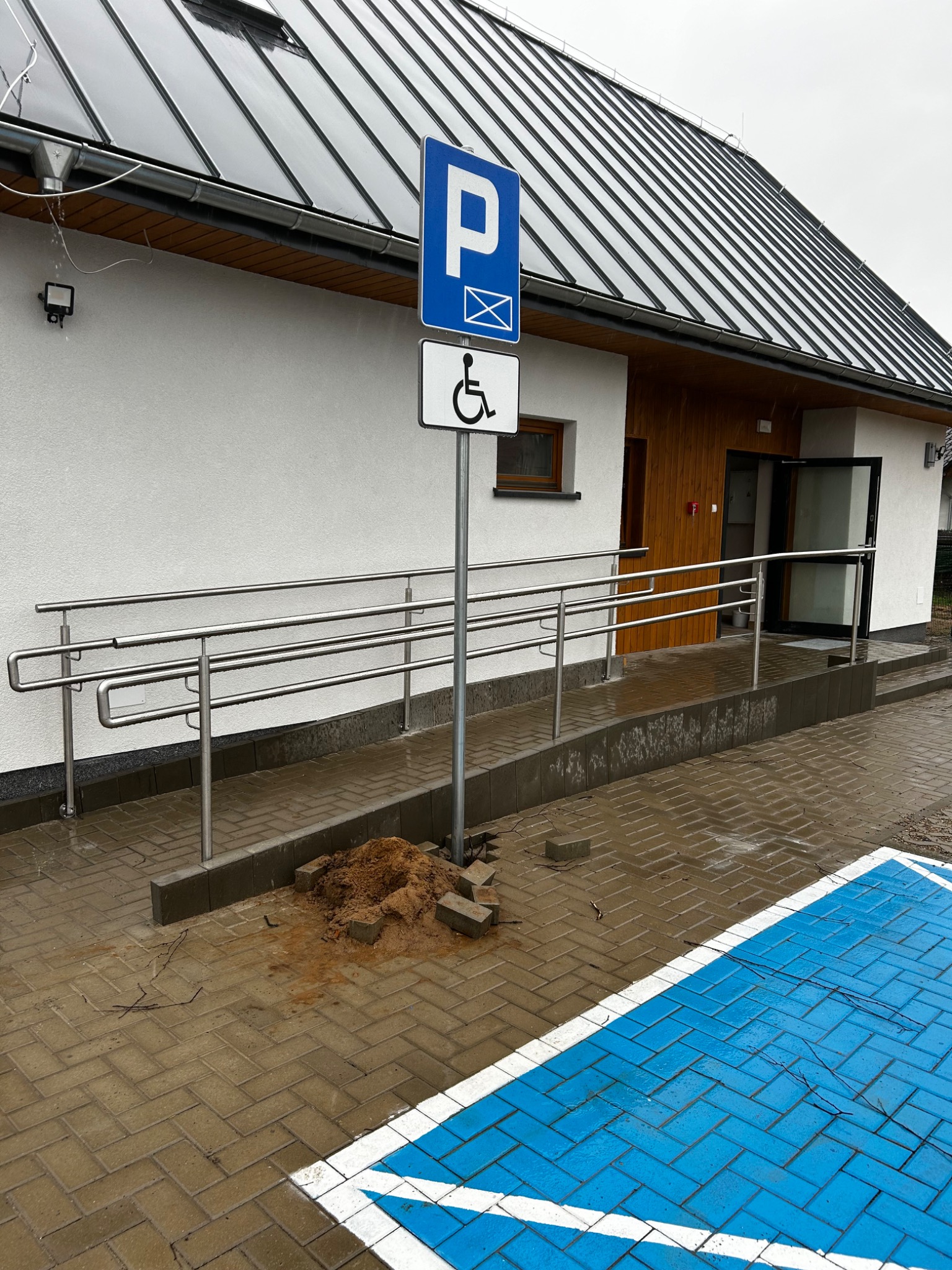 Zewnętrzna rampa dla niepełnosprawnych z nierdzewnymi balustradami prowadząca do budynku z drewnianymi elementami, obok znak parkingu dla niepełnosprawnych, mokra kostka brukowa.