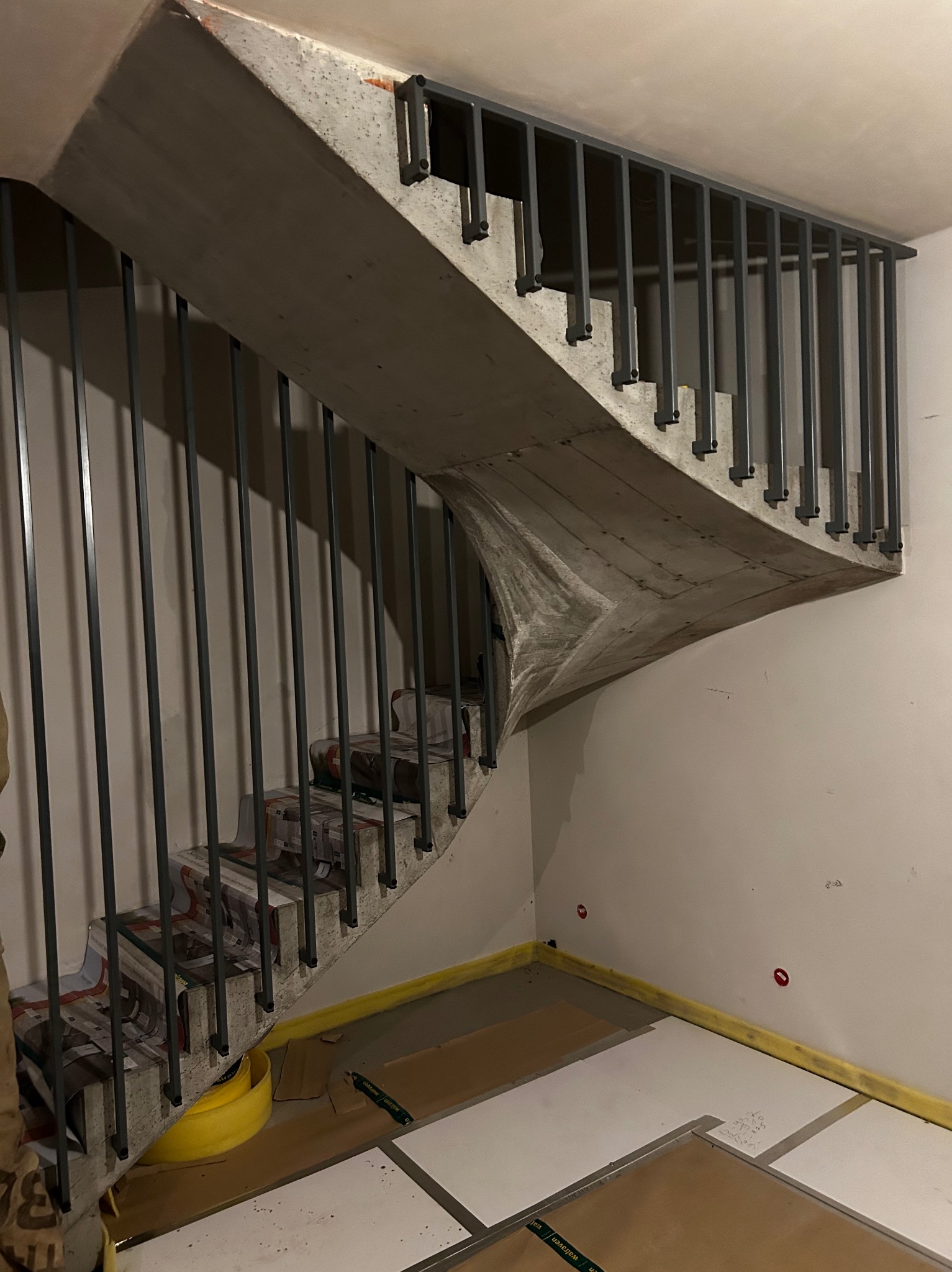 Surowe, betonowe schody z metalową balustradą w trakcie prac wykończeniowych, zabezpieczone kartonem i folią malarską.