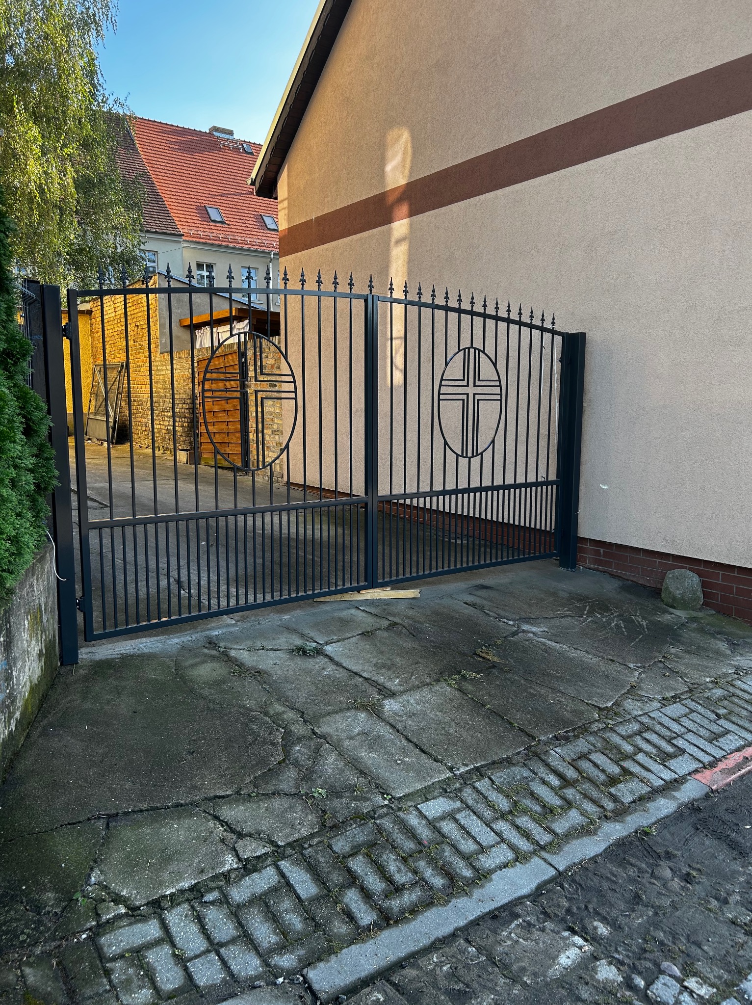 Czarna, metalowa brama wjazdowa z symbolem krzyża w okręgu, prowadząca na posesję z budynkiem o beżowej elewacji i czerwonym dachu, widoczna kostka brukowa i fragment zieleni.