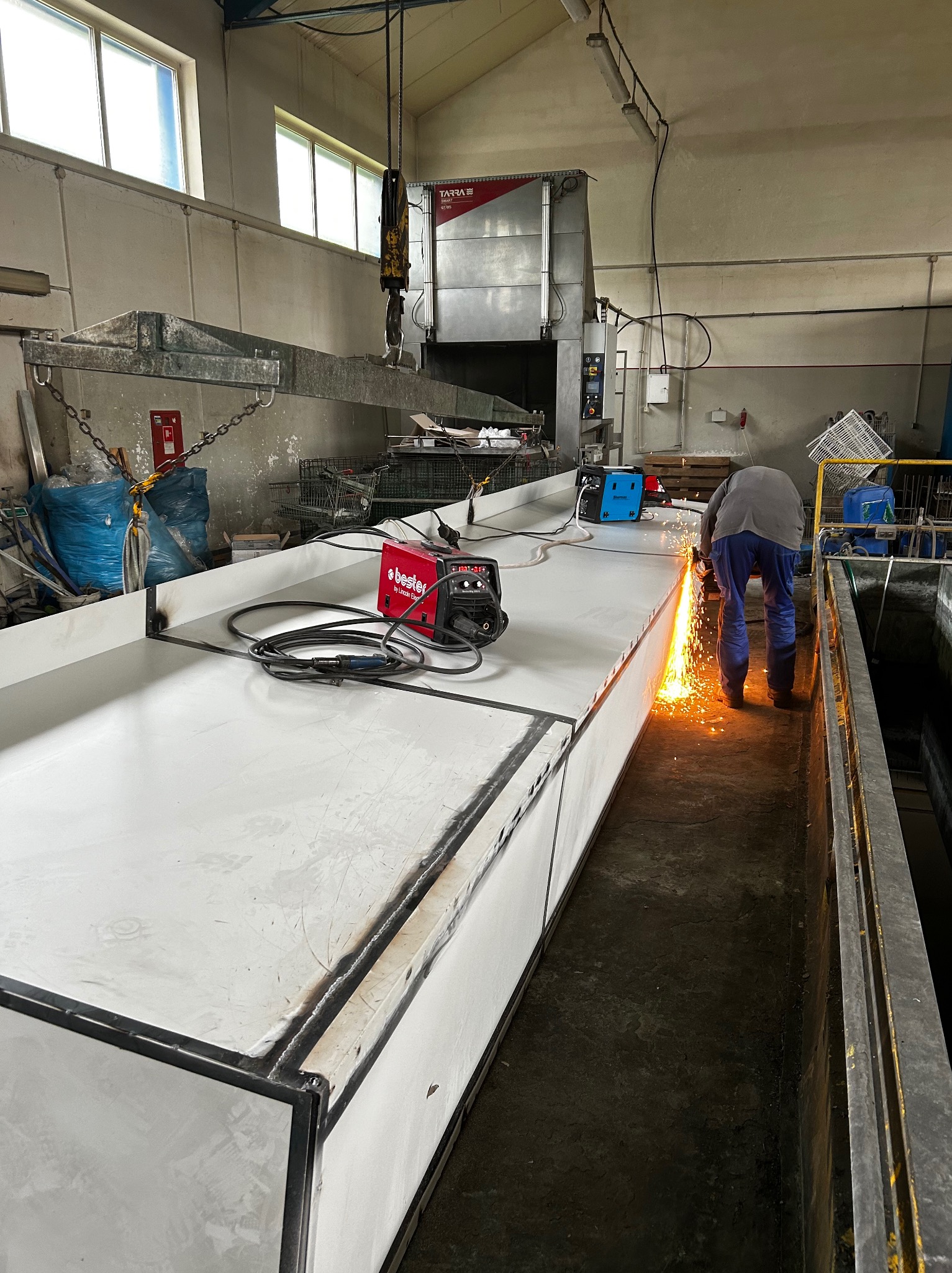 Pracownik w warsztacie szlifuje spaw na dużej, metalowej konstrukcji, iskry lecą, widoczny sprzęt spawalniczy i suwnica pod sufitem.