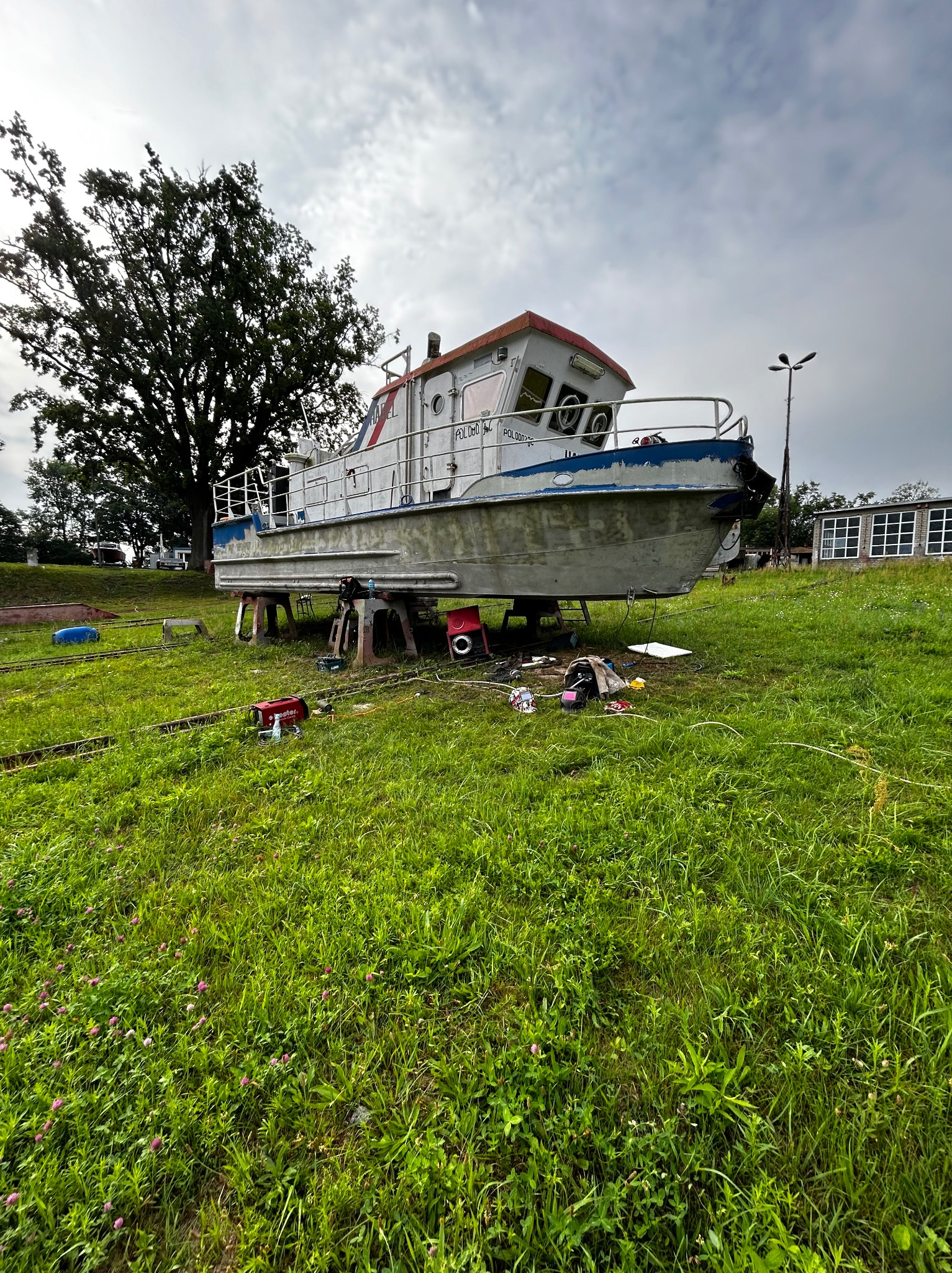 Łódź motorowa na metalowych podporach, prace konserwacyjne na trawiastym terenie, widoczne narzędzia i spawarka, pochmurne niebo w tle.