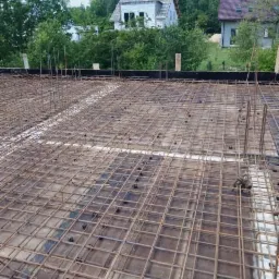 Siatka zbrojeniowa na deskowaniu, przygotowana do wylania betonu, z widocznymi elementami dystansowymi, na tle domów w otoczeniu zieleni.