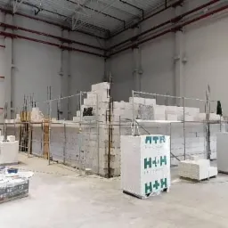 Wnętrze hali produkcyjnej z wznoszoną ścianą z bloczków H+H na rusztowaniach, widoczne czerwone rury instalacji pod stropem i pracownicy w odblaskowych kamizelkach.