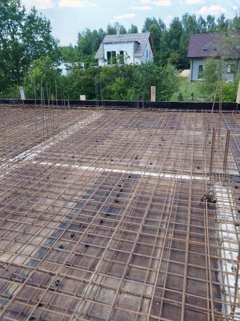 Siatka zbrojeniowa na deskowaniu, przygotowana do wylania betonu, z widocznymi elementami dystansowymi, na tle domów w otoczeniu zieleni.