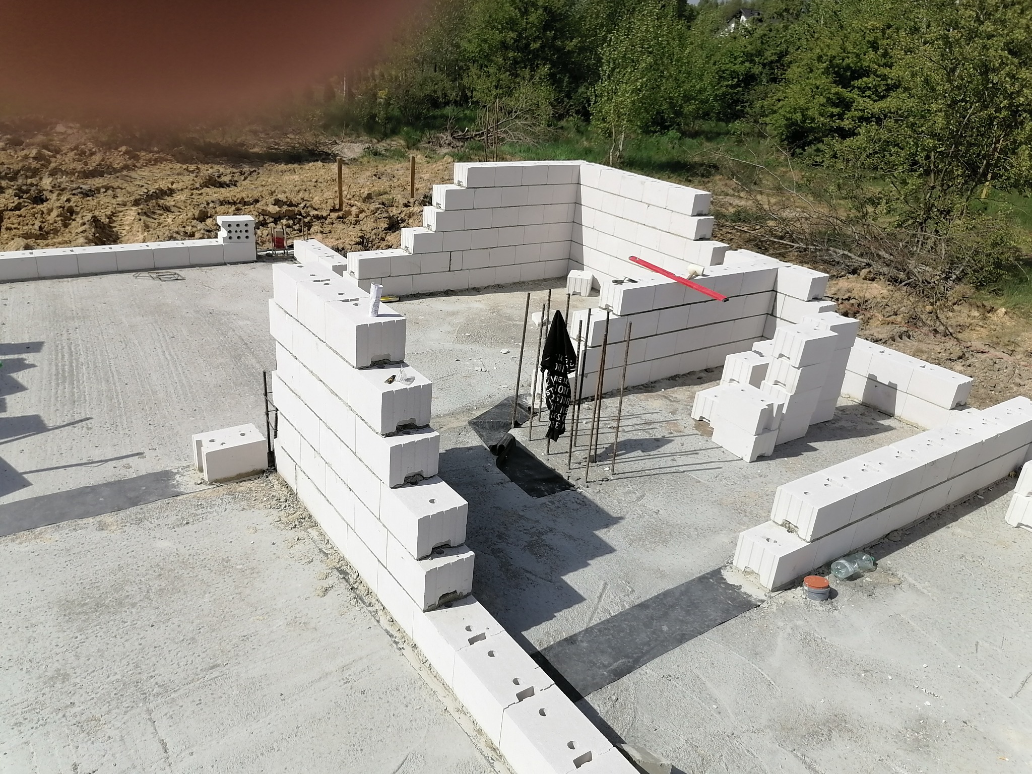 Wznoszenie ścian parteru z białych bloczków na fundamencie, widoczne zbrojenia i poziomica, w tle zieleń.