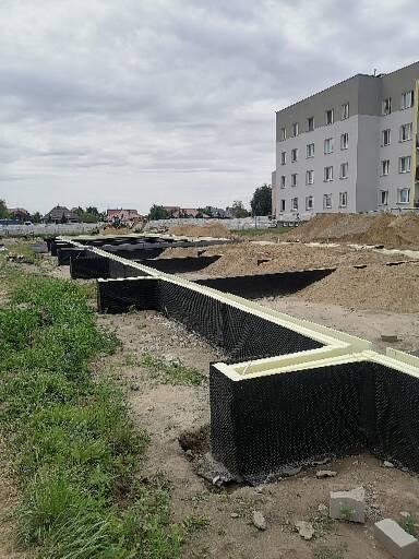 Formowanie szalunków traconych z czarnego tworzywa sztucznego z białym wypełnieniem, tworzące fundamenty pod budynek wielorodzinny w trakcie budowy, widoczne wykopane rowy, ziemia i budynek w tle.