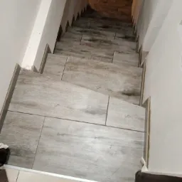 Schody wyłożone szarymi płytkami imitującymi drewno, widok z góry, zarys białej ściany po obu stronach.