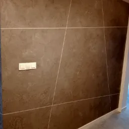 Ściana z dekoracyjną, brązową gładzią strukturalną, podzielona białymi listwami tworzącymi geometryczny wzór. Widoczny włącznik światła z trzema przyciskami oraz biała listwa przypodłogowa.