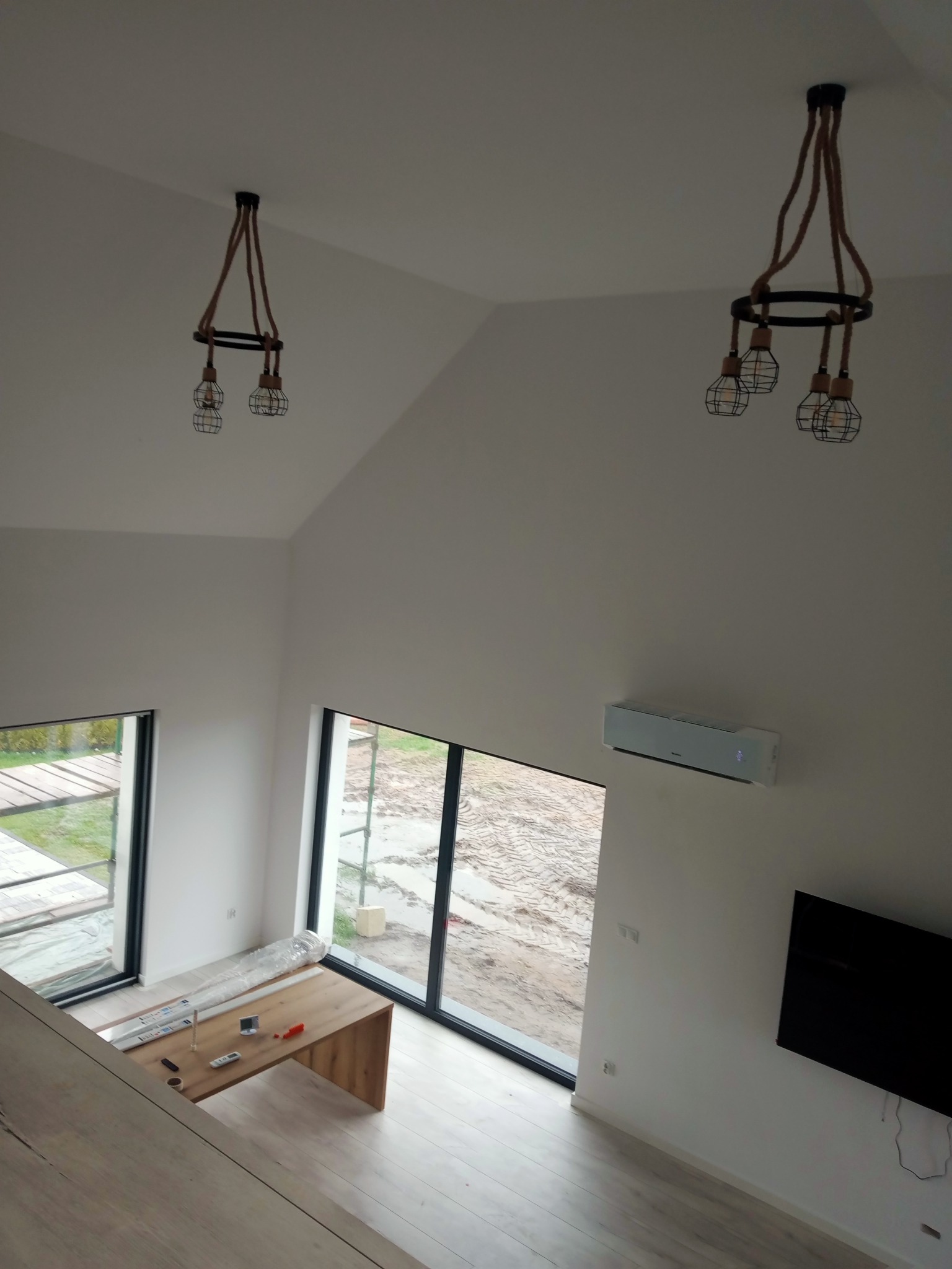 Wysoki, jasny pokój z dwoma oknami, klimatyzacją i dwoma industrialnymi lampami wiszącymi z brązowymi sznurami, widok z góry na drewniany blat stołu z narzędziami i materiałami budowlanymi.