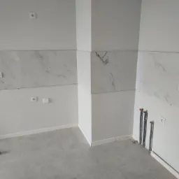 Ściana wyłożona białymi płytami imitującymi marmur, z widocznymi gniazdkami elektrycznymi i wystającymi rurami instalacji wodnej, szare płytki podłogowe, listwy przypodłogowe