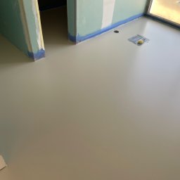 GS EPOXYFLOORS S.C. - Jasnoszara, gładka powierzchnia w nowym wnętrzu z zabezpieczonymi ścianami i oknami, sugerująca świeżo wylaną, jednolitą podłogę. Widoczny otwór w podłodze.