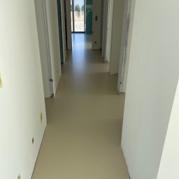 GS EPOXYFLOORS S.C. - Beżowa, gładka podłoga w korytarzu z białymi ścianami i otworami drzwiowymi. Widok w głąb pomieszczenia z oknem na końcu. Minimalistyczny design, świeżo wylana powierzchnia.