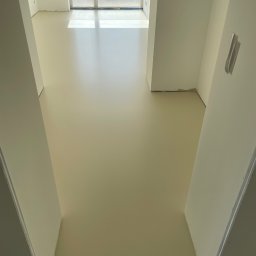 GS EPOXYFLOORS S.C. - Jasna, gładka podłoga w nowym wnętrzu, widziana z góry. Świeżo wylana, beżowa powierzchnia odbija światło z okna. Przy ścianach widoczne zabezpieczenia.