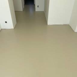GS EPOXYFLOORS S.C. - Beżowa, gładka podłoga w jasnym wnętrzu z białymi ścianami i filarami. Widok na korytarz w głębi. Gniazdko elektryczne na ścianie.