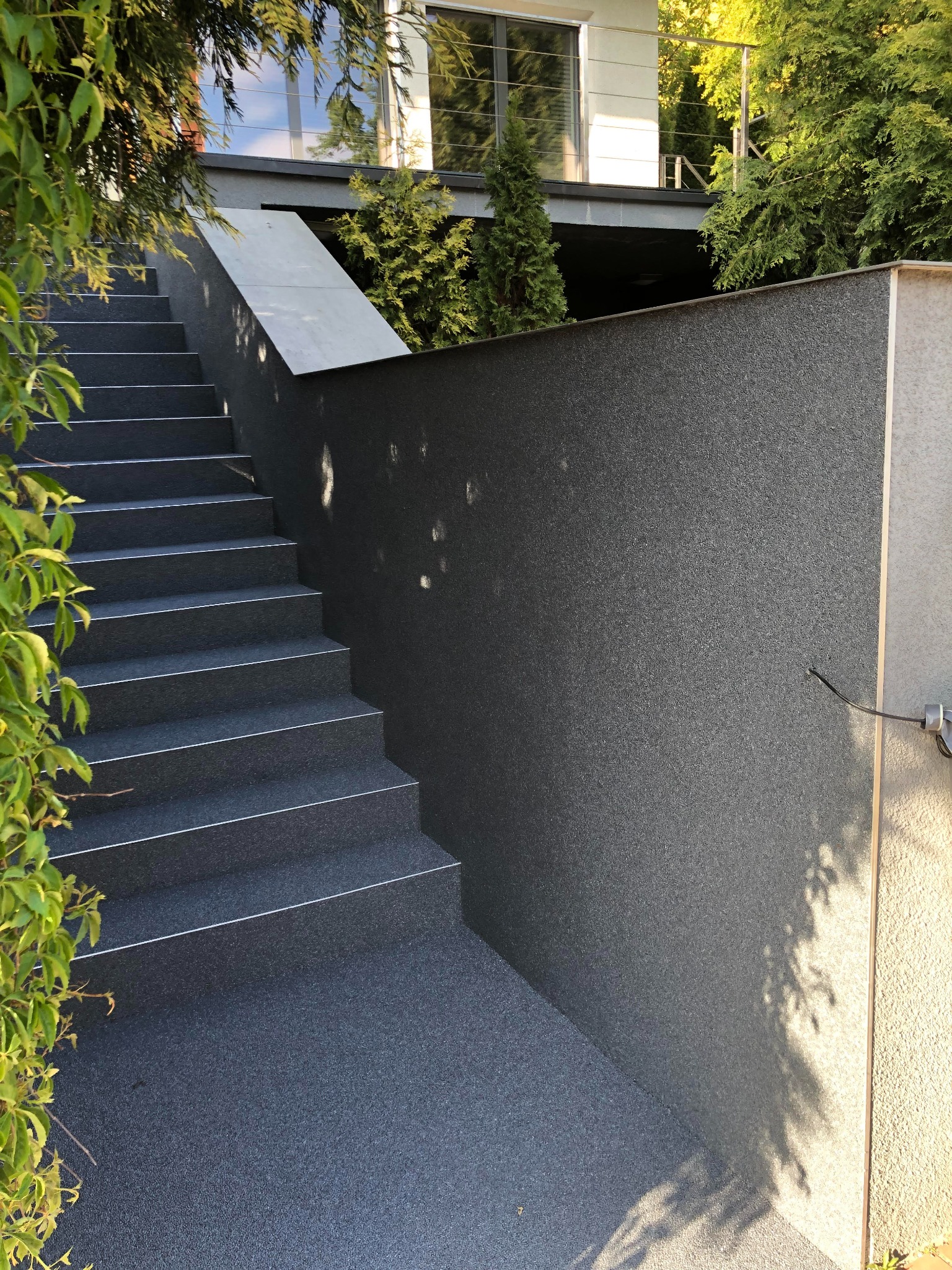 Zewnętrzne schody pokryte ciemną, drobnoziarnistą nawierzchnią, z białymi krawędziami, prowadzące w górę wzdłuż ciemnoszarej ściany, z zieloną roślinnością po lewej stronie i nowoczesnym budynkiem...