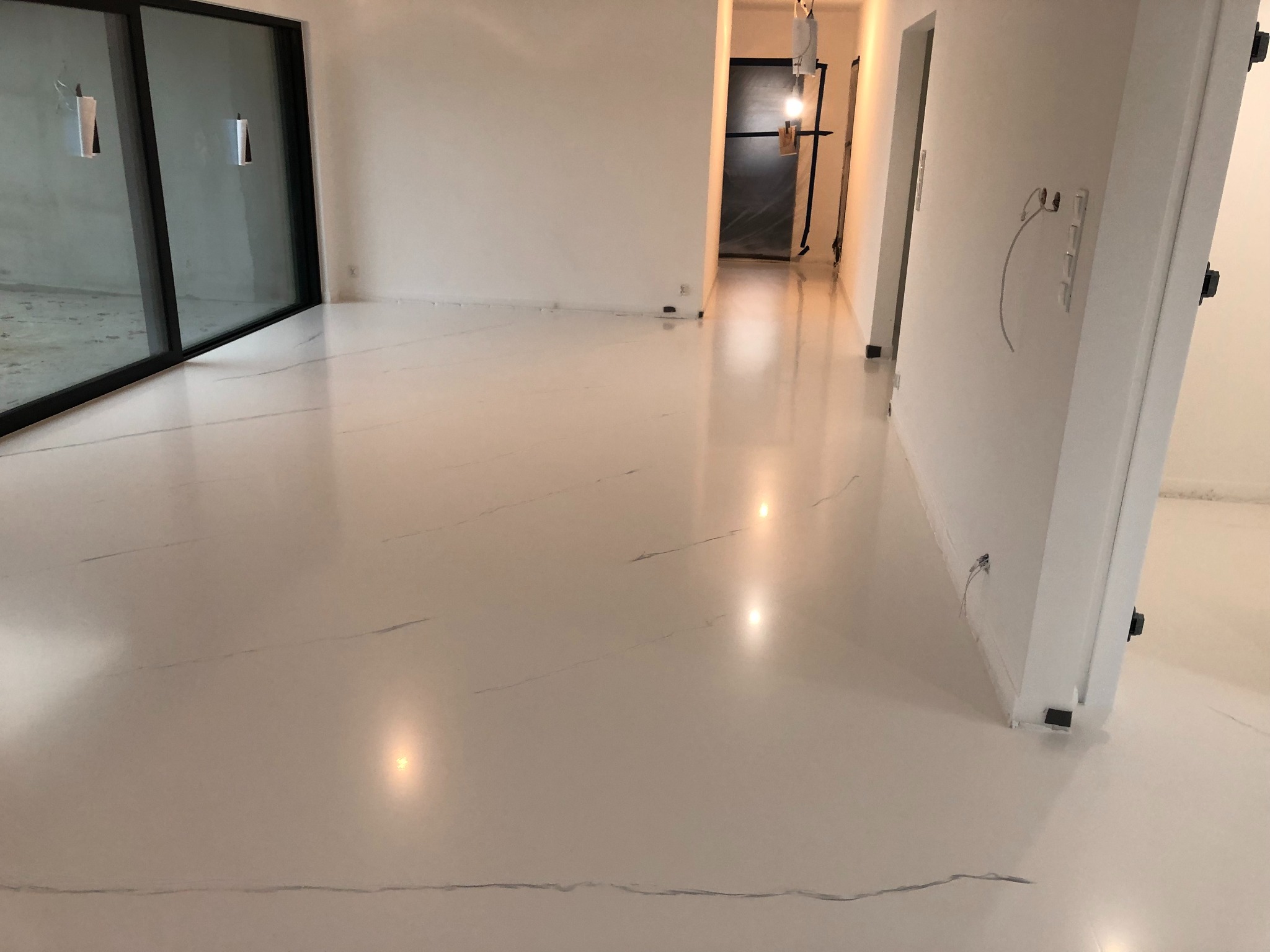 Biała posadzka żywiczna z delikatnym marmurkowym wzorem w przestronnym, jasnym wnętrzu z dużym oknem i przygotowanymi instalacjami elektrycznymi.