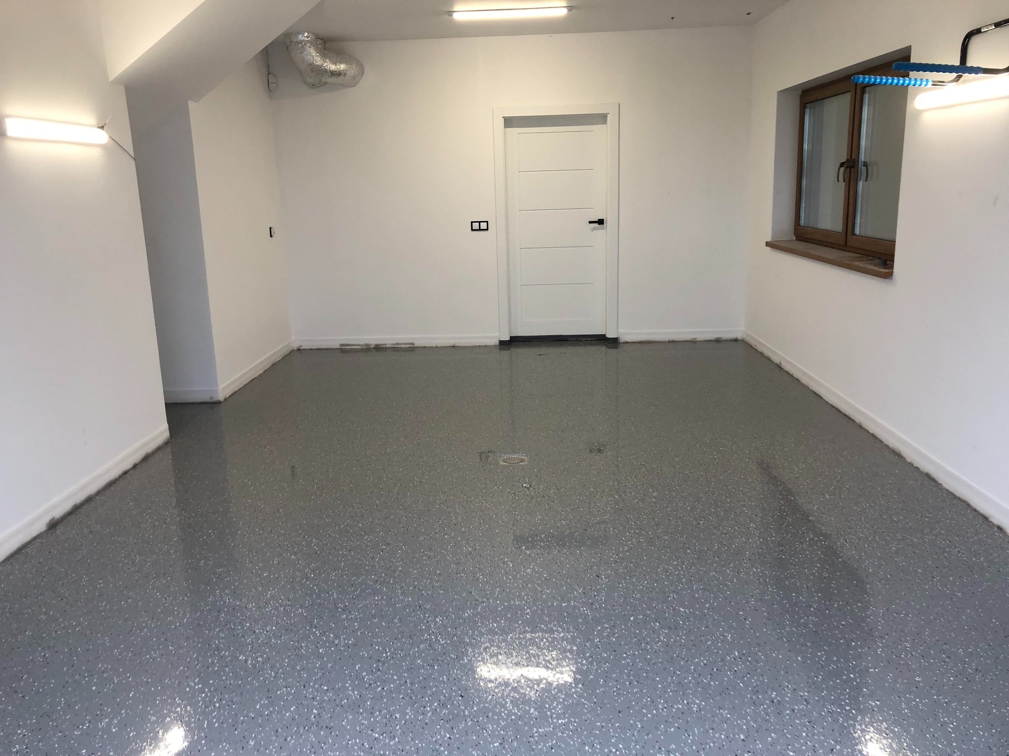 Szara, błyszcząca podłoga z drobnymi, kolorowymi wtrąceniami w nowym, jasnym pomieszczeniu z białą ścianą, drzwiami i oknem.