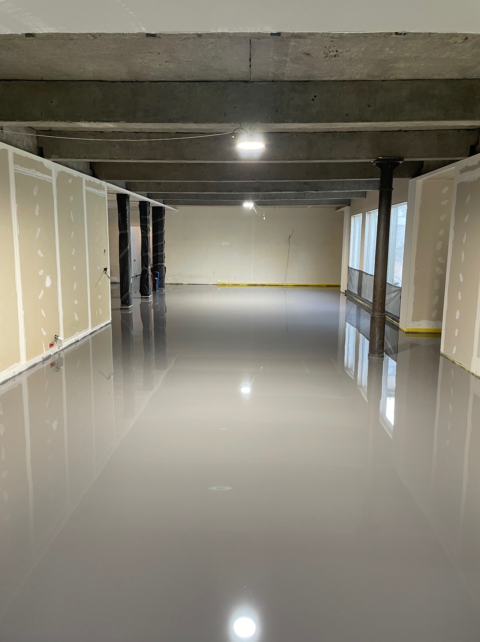 Błyszcząca, szara posadzka w surowym wnętrzu z betonowym stropem i ścianami przygotowanymi do malowania. Widoczne słupy nośne i okna z żółtą folią zabezpieczającą.