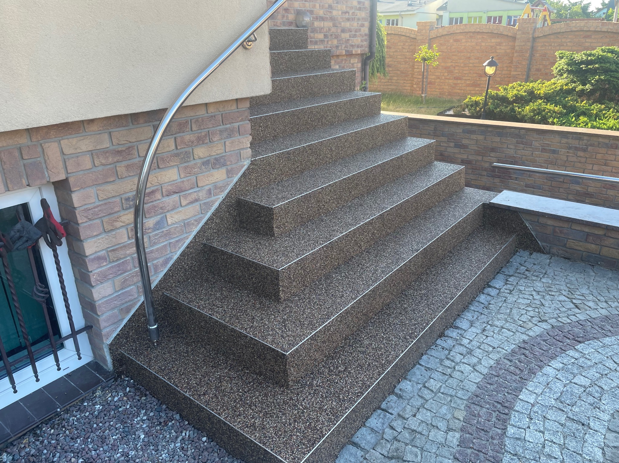Schody zewnętrzne z posadzką żywiczną w kolorze brązowym, wykończone metalowymi listwami. Widoczna barierka ze stali nierdzewnej i kostka brukowa na podwórku.