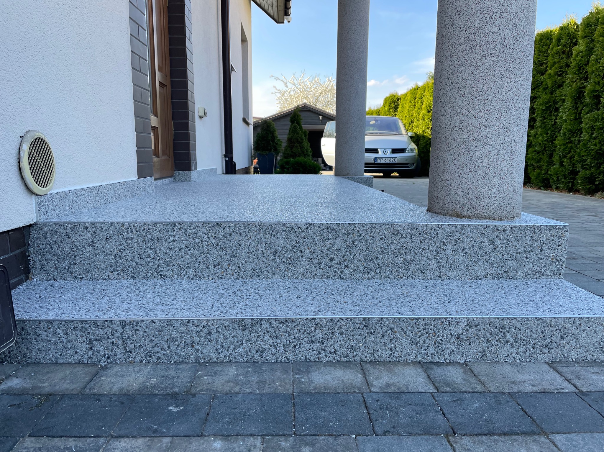 Schody wejściowe z szarej, drobnoziarnistej posadzki żywicznej, wykończone metalowymi listwami. W tle elewacja domu i zaparkowany samochód.