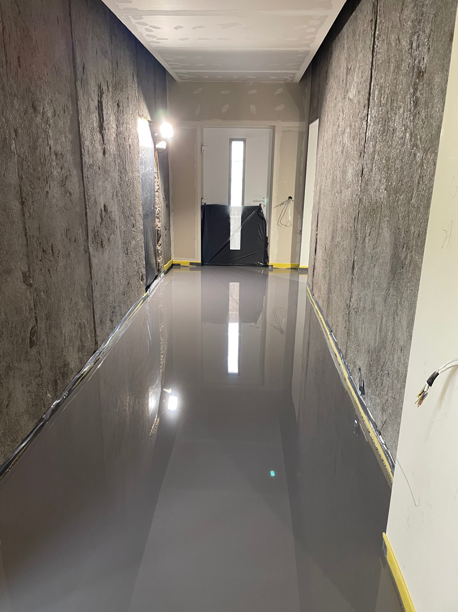 Świeżo wylana, szara podłoga w korytarzu, zabezpieczona folią i taśmą malarską przy ścianach o surowym wykończeniu. Widoczne kable i lampa halogenowa.