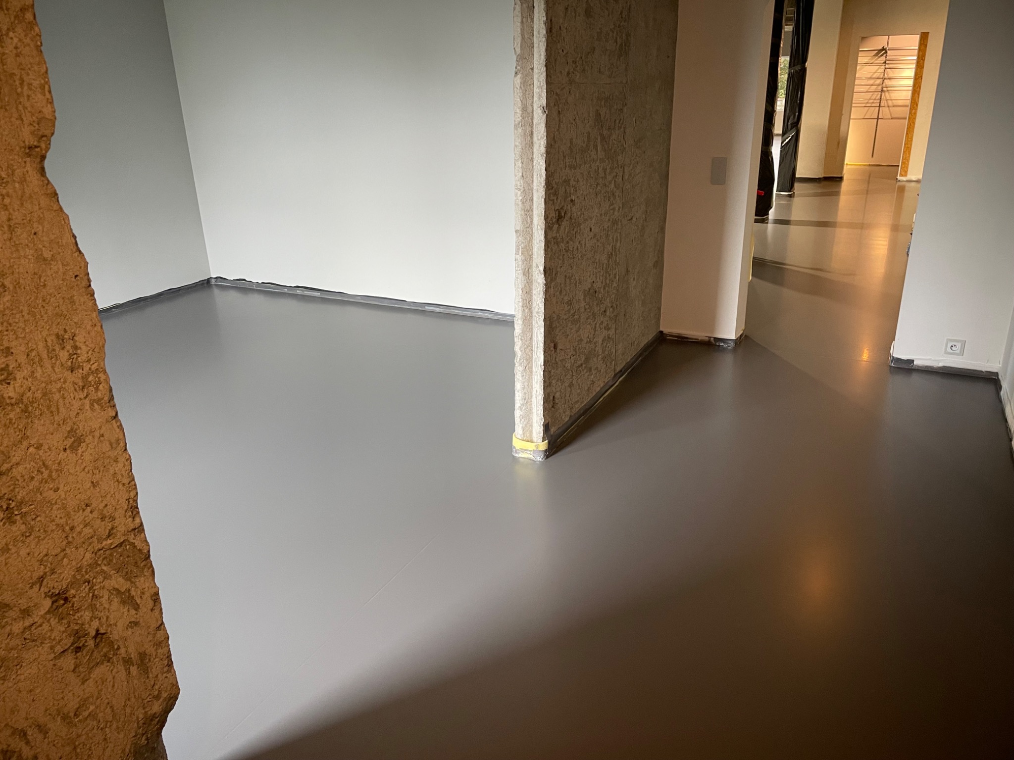 Szara, gładka posadzka w surowym wnętrzu z betonowym filarem. Widok na sąsiednie pomieszczenie z podobną podłogą. Minimalistyczny design, świeżo wylana powierzchnia.