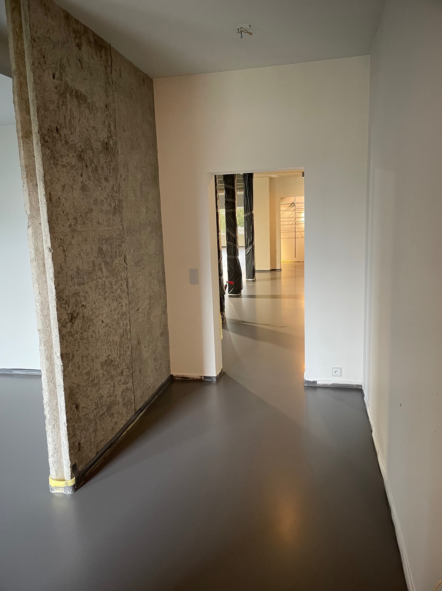 Szara, gładka posadzka w jasnym wnętrzu z surowym betonem. Widok na kolejne pomieszczenie przez otwarte przejście. Minimalistyczny design, świeżo wylana podłoga.