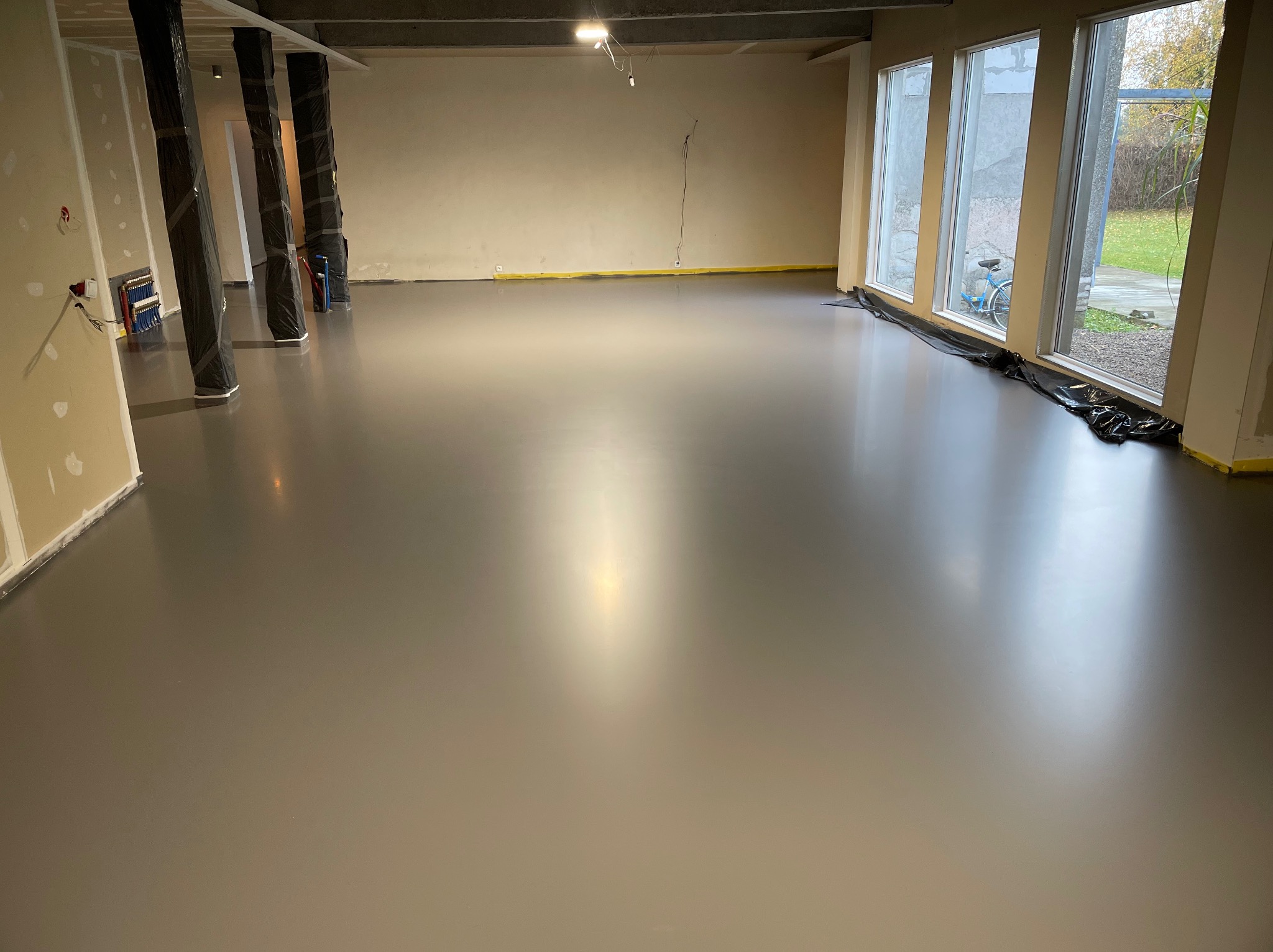 Szara, gładka posadzka w jasnym pomieszczeniu z oknami i kolumnami zabezpieczonymi folią. Żółta taśma malarska wzdłuż ścian. Widok na zewnątrz przez okna.
