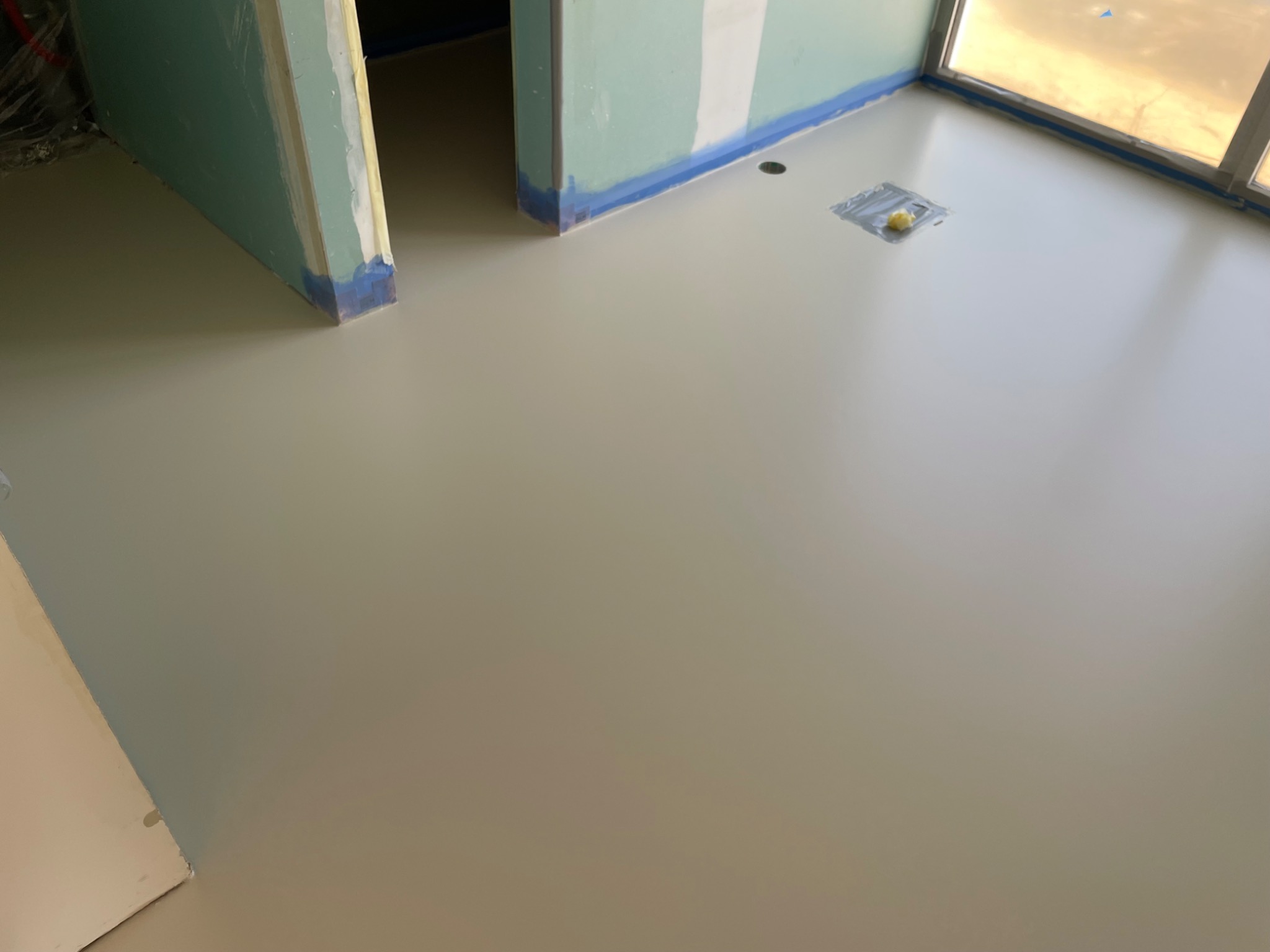 Jasnoszara, gładka powierzchnia w nowym wnętrzu z zabezpieczonymi ścianami i oknami, sugerująca świeżo wylaną, jednolitą podłogę. Widoczny otwór w podłodze.