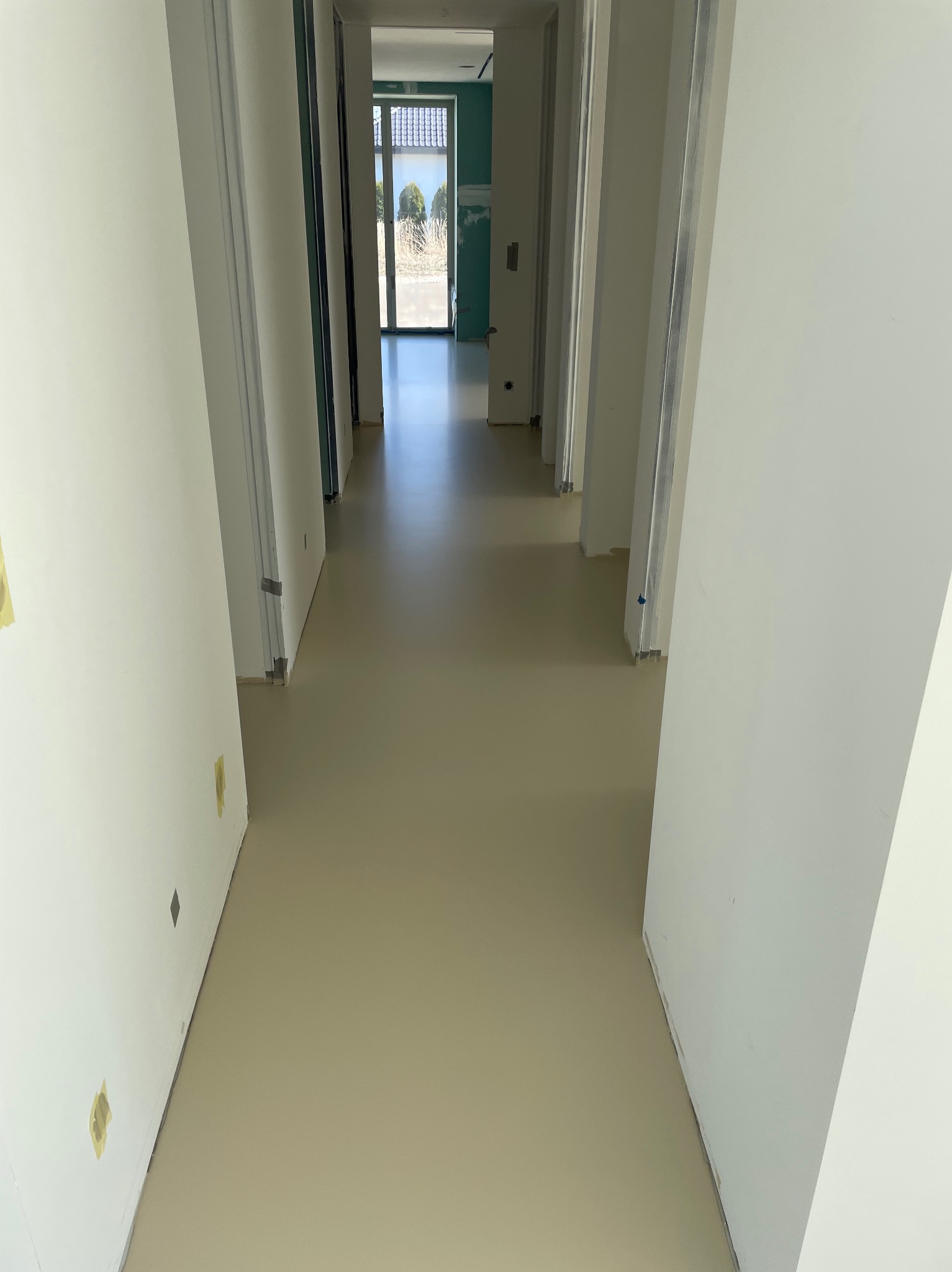 Beżowa, gładka podłoga w korytarzu z białymi ścianami i otworami drzwiowymi. Widok w głąb pomieszczenia z oknem na końcu. Minimalistyczny design, świeżo wylana powierzchnia.