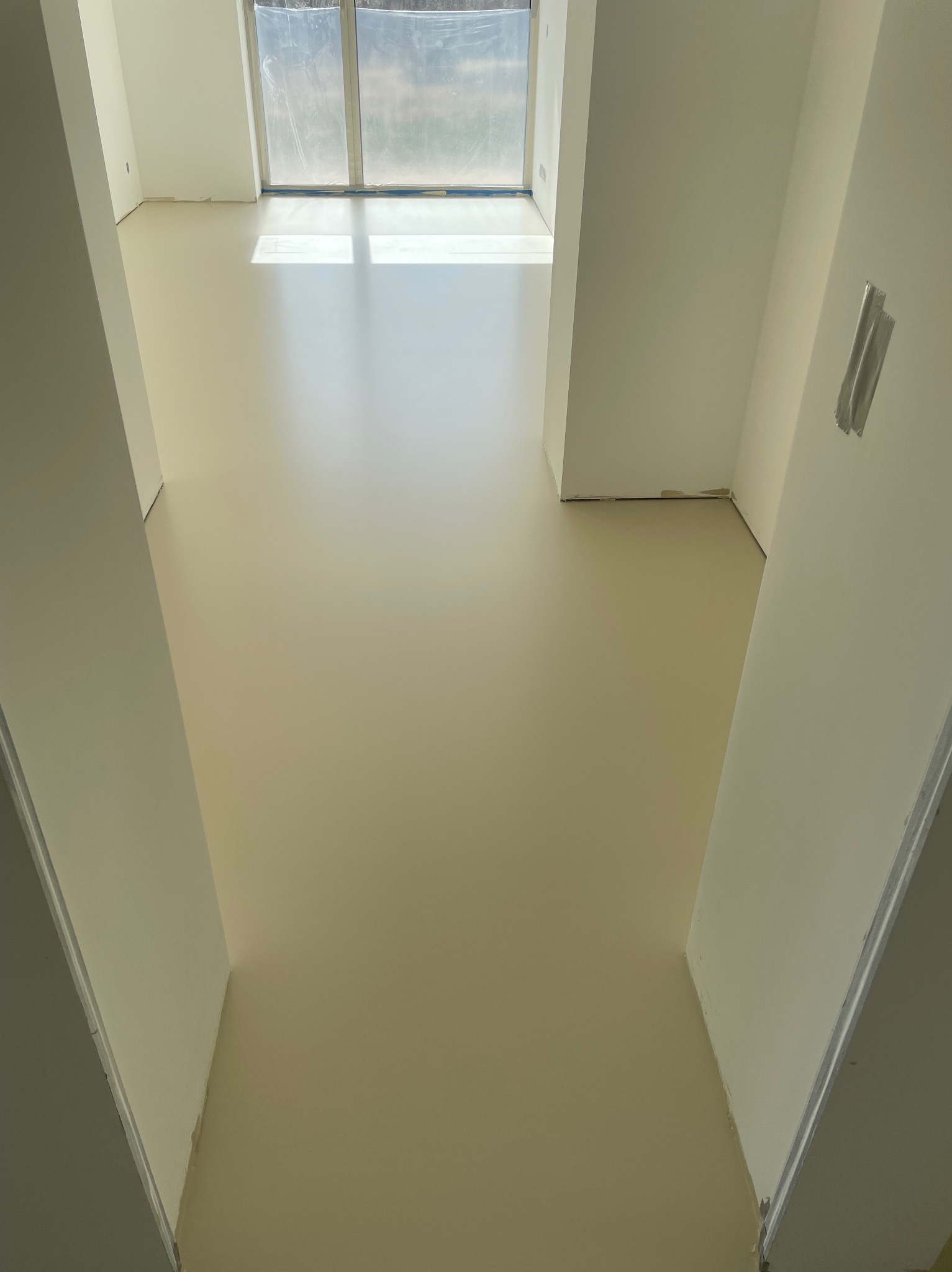 Jasna, gładka podłoga w nowym wnętrzu, widziana z góry. Świeżo wylana, beżowa powierzchnia odbija światło z okna. Przy ścianach widoczne zabezpieczenia.