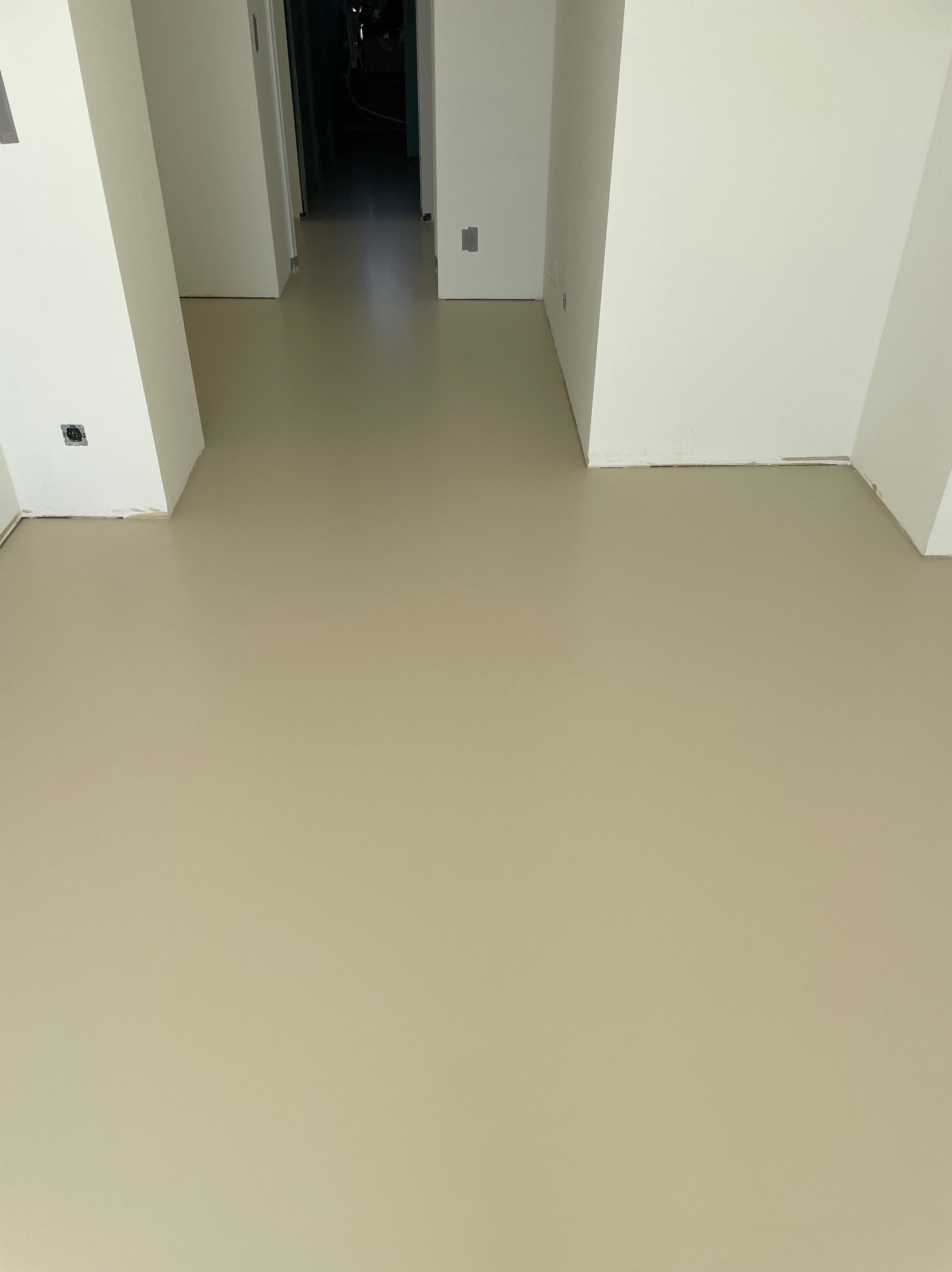 Beżowa, gładka podłoga w jasnym wnętrzu z białymi ścianami i filarami. Widok na korytarz w głębi. Gniazdko elektryczne na ścianie.
