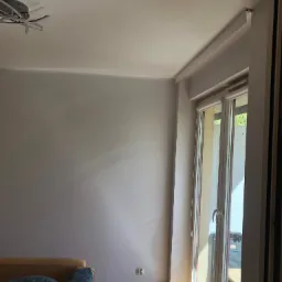 Pokój z szarą ścianą, drewnianą podłogą, oknem balkonowym z roletami i nowoczesną lampą sufitową