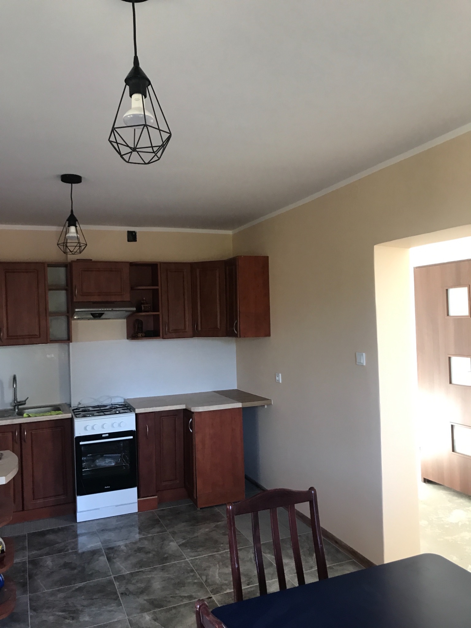 Ukończona kuchnia z brązowymi szafkami, białymi płytkami nad blatem, nowoczesnymi lampami wiszącymi z czarnymi kloszami i ciemną podłogą z płytek.