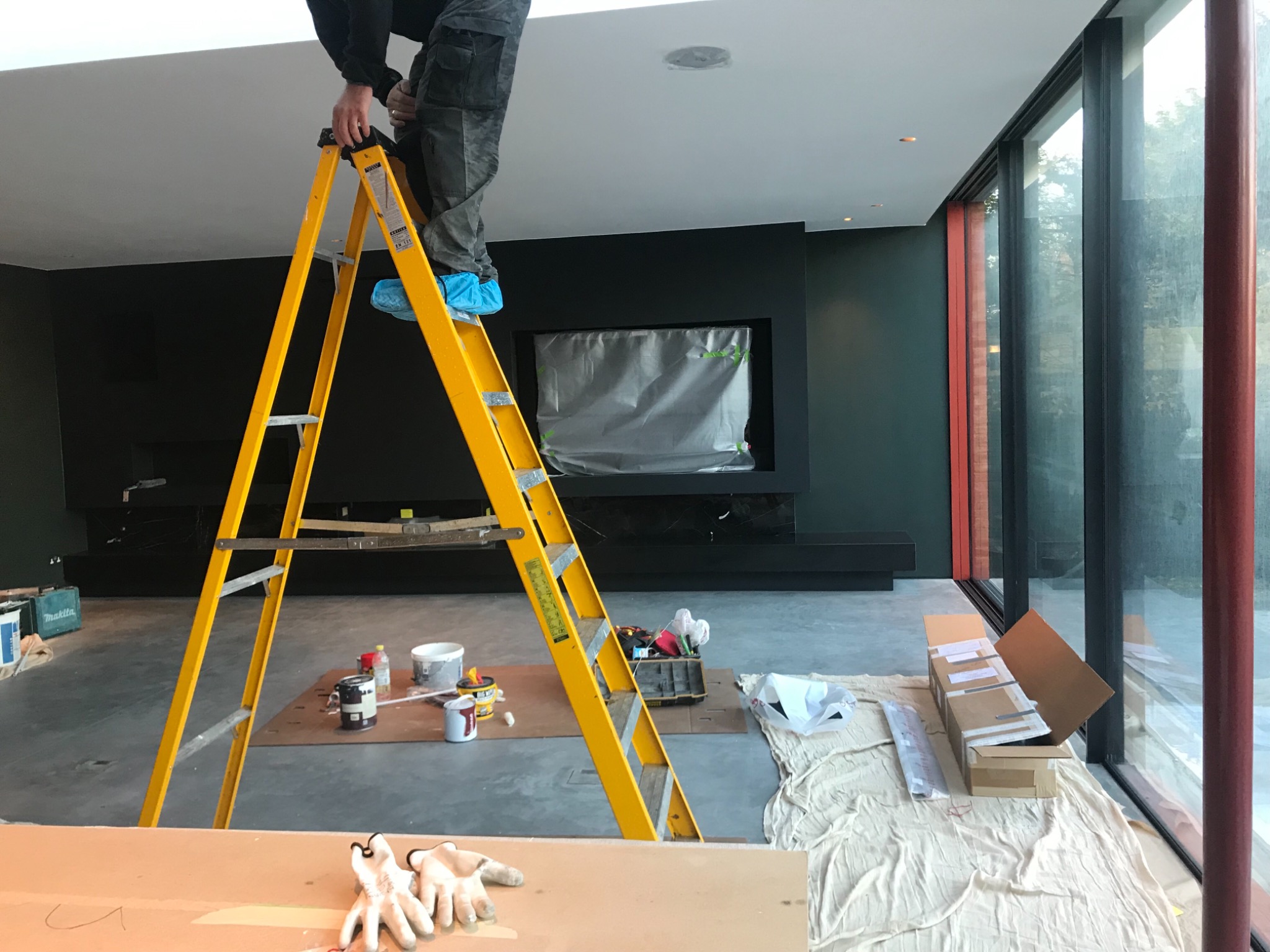 Malarz na żółtej drabinie, w niebieskich ochraniaczach na obuwiu, przygotowuje się do malowania sufitu w nowoczesnym salonie z dużymi oknami i ciemnozieloną ścianą z kominkiem zabezpieczonym folią.