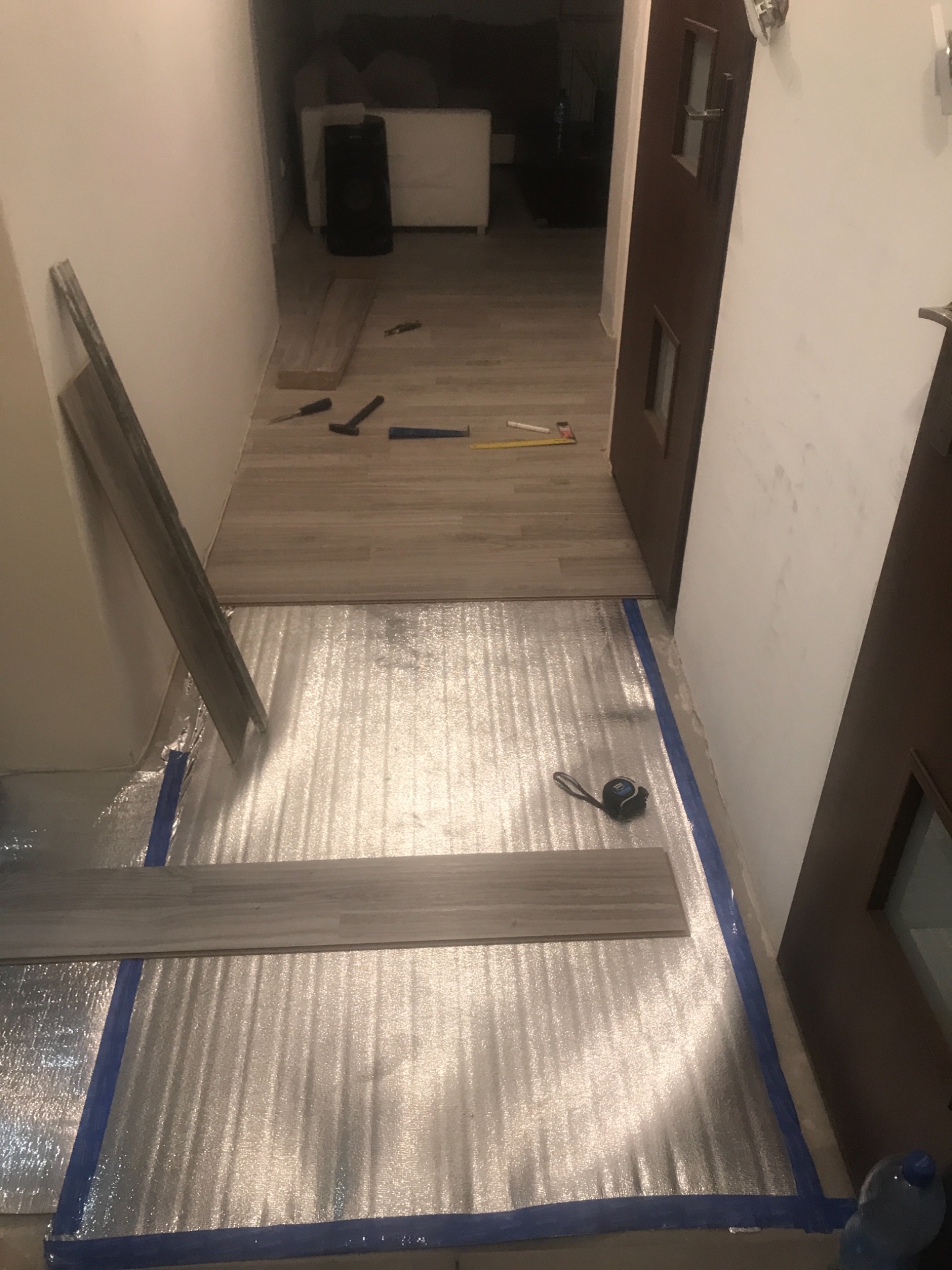 Proces montażu paneli podłogowych w korytarzu, widoczna folia izolacyjna, narzędzia, przymierzone panele i fragment ułożonej podłogi.