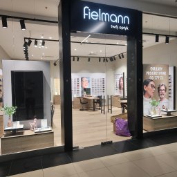 SD-UNION Dariusz Stachowiak - Wnętrze salonu optycznego Fielmann z ekspozycją okularów, oświetleniem punktowym i elementami dekoracyjnymi, widoczne plakaty reklamowe i purpurowa torba przy wejściu.