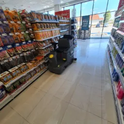 Automat do mycia podłóg w supermarkecie, czyszczący kafelki pomiędzy półkami z towarem, widoczne ślady wilgoci po myciu.