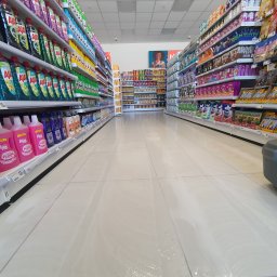 SD-UNION Dariusz Stachowiak - Maszyna czyszcząca myje podłogę w supermarkecie, widok na alejkę z półkami pełnymi środków czystości i artykułów spożywczych, mokra podłoga odbija światło.