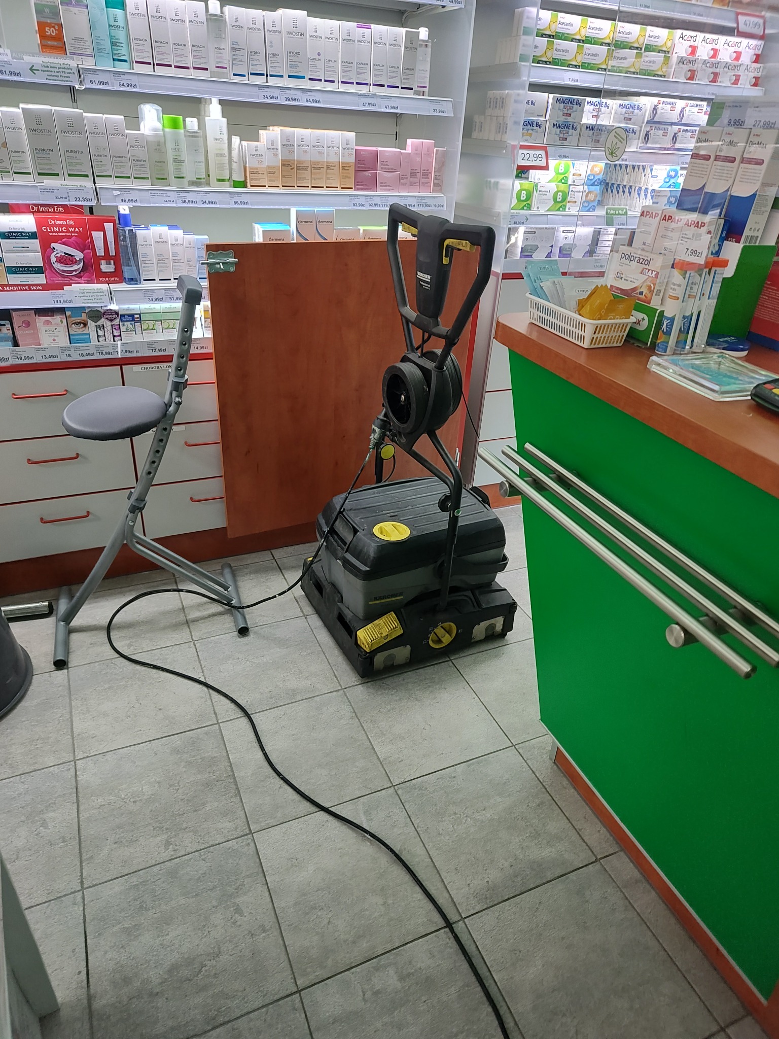 Automat szorująco-zbierający Kärcher BR 30/4 C w aptece, stojący obok krzesła regulowanego i zielonej lady z ekspozycją leków i kosmetyków w tle.