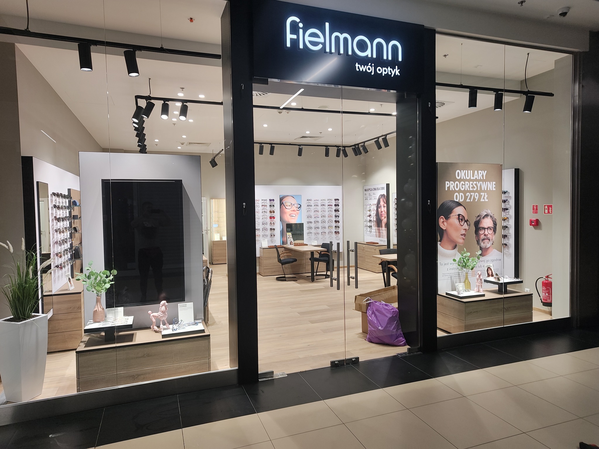 Wnętrze salonu optycznego Fielmann z ekspozycją okularów, oświetleniem punktowym i elementami dekoracyjnymi, widoczne plakaty reklamowe i purpurowa torba przy wejściu.