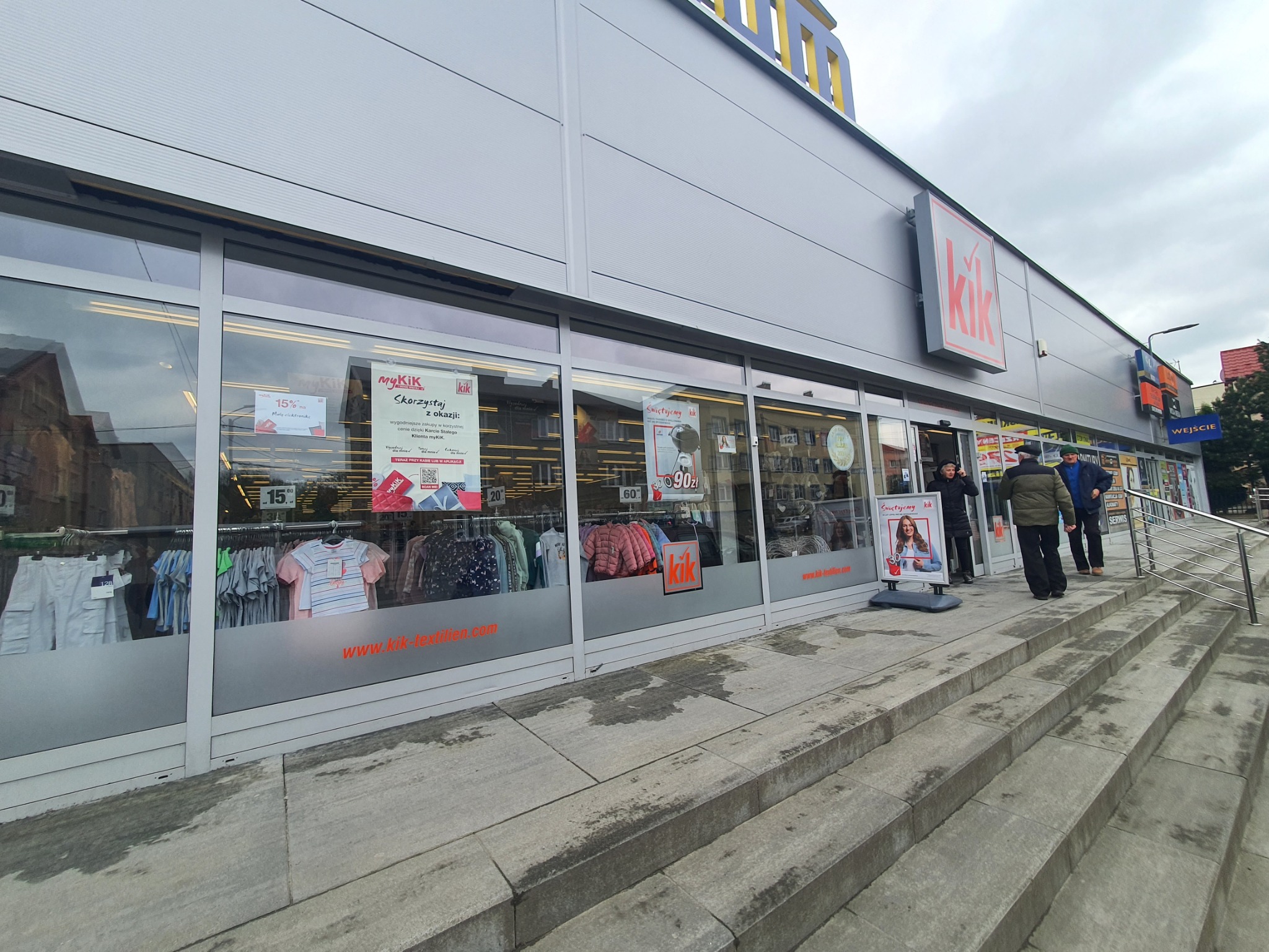 Widok zewnętrzny sklepu KiK z odzieżą i artykułami gospodarstwa domowego, z widocznymi witrynami wystawowymi, schodami wejściowymi i szyldem z logo marki.