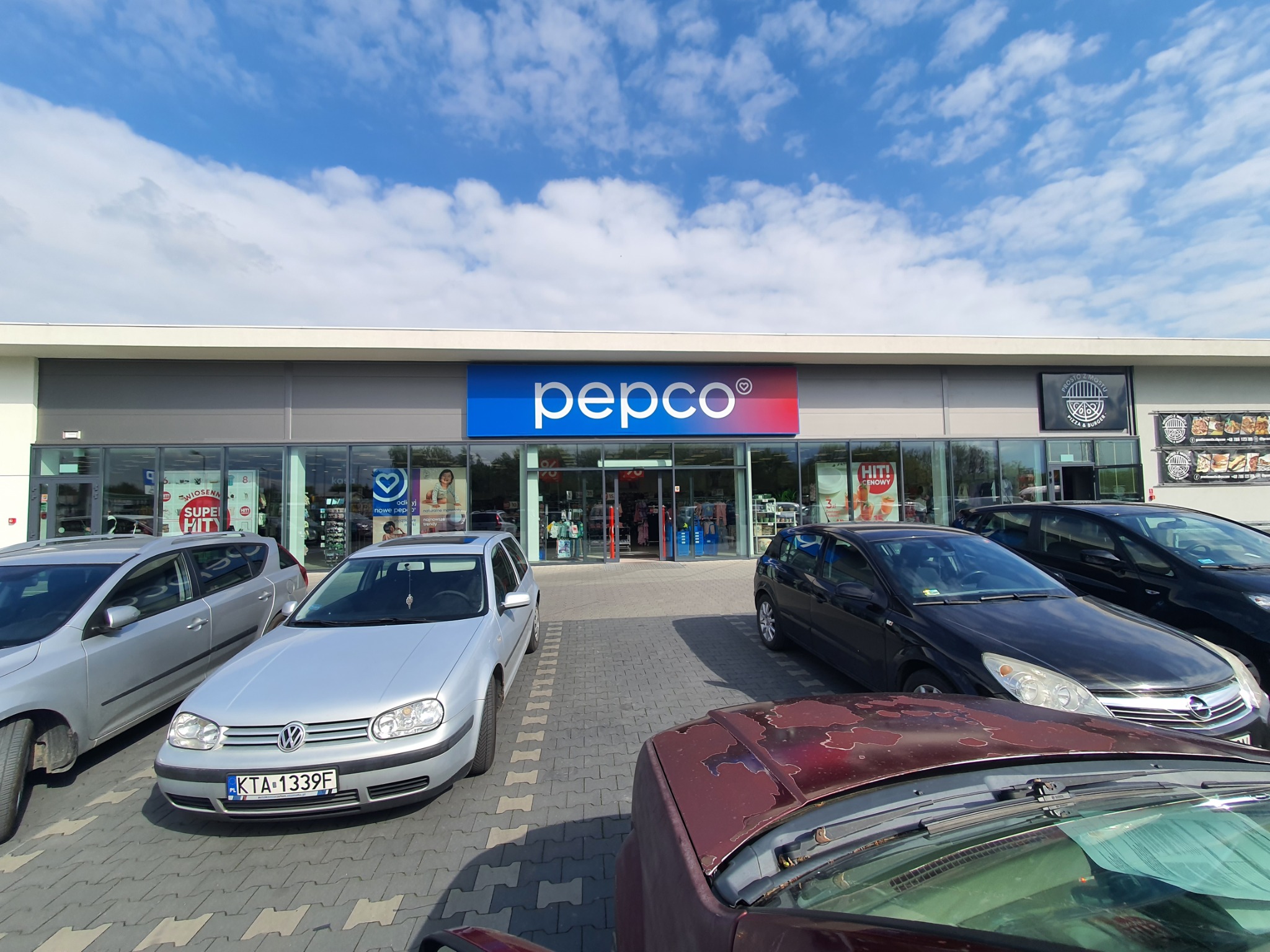 Fasada sklepu Pepco z zaparkowanymi samochodami na pierwszym planie, widok na wejście i szyld z logo na tle błękitnego nieba z chmurami.