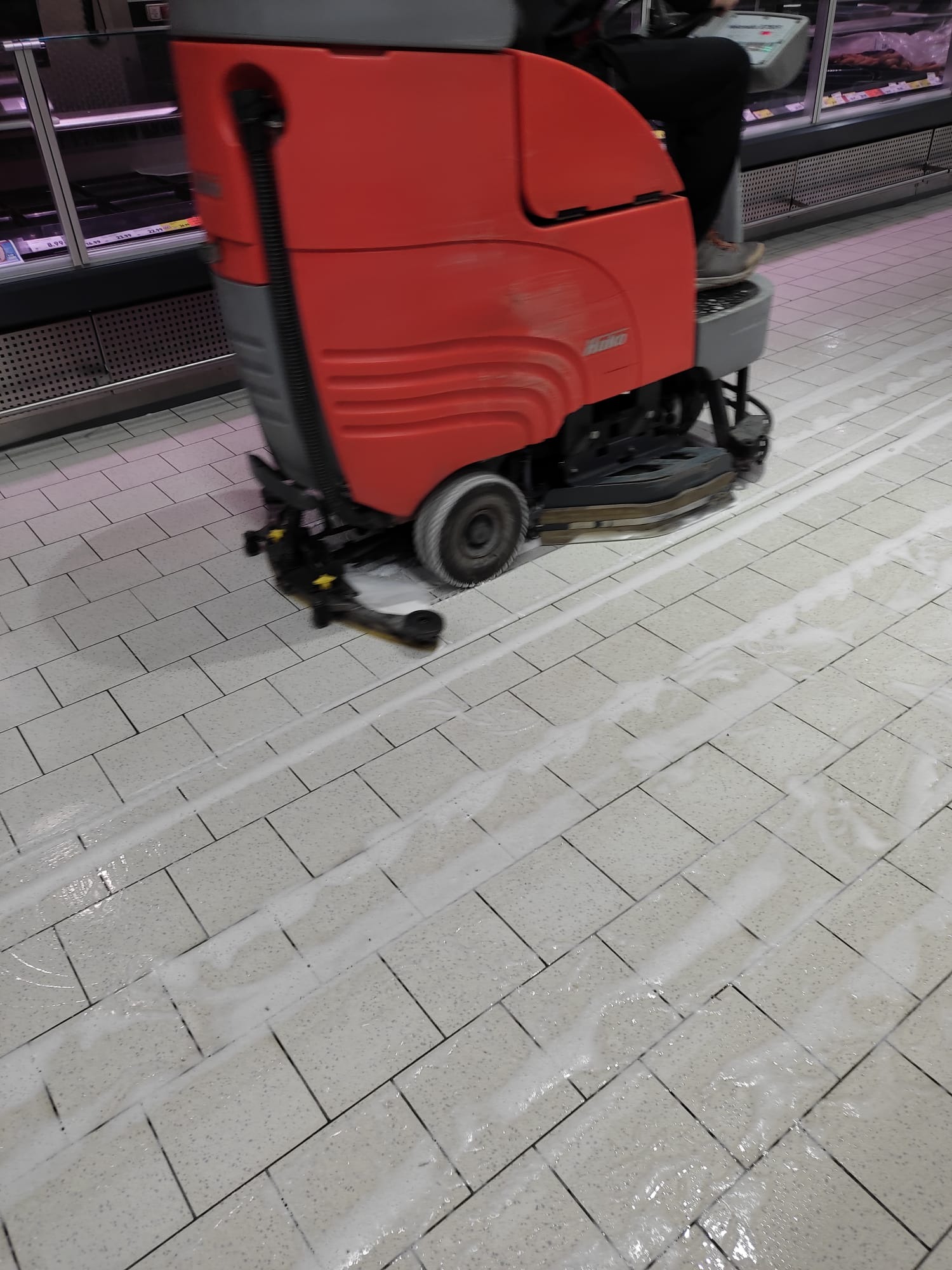 Czerwona maszyna czyszcząca z operatorem w trakcie pracy na jasnej, kafelkowanej podłodze w sklepie, pozostawiająca za sobą mokre ślady.