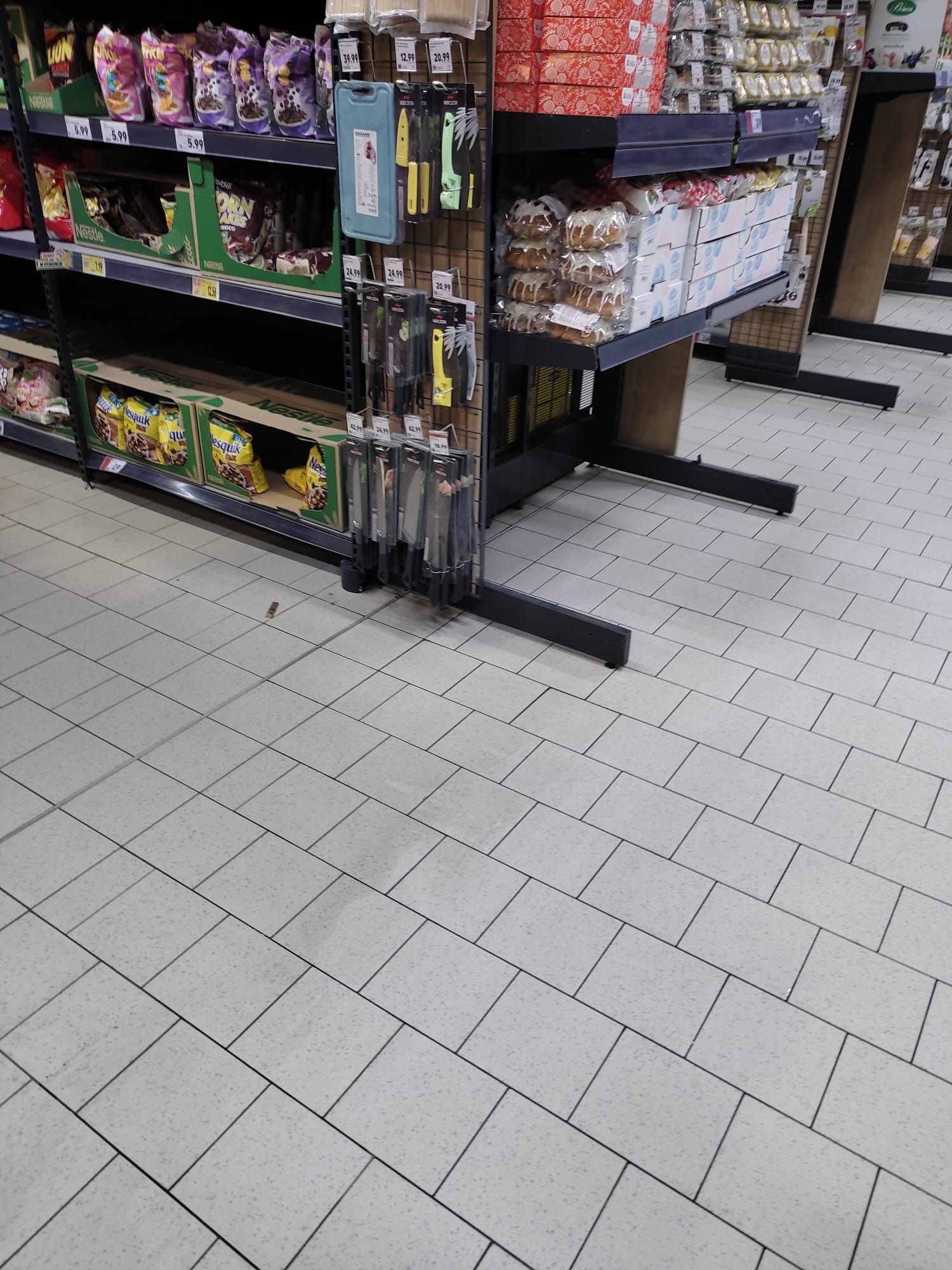 Widok fragmentu sklepu z półkami pełnymi różnorodnych produktów spożywczych i akcesoriów kuchennych, z przewagą płytek podłogowych w neutralnym odcieniu.